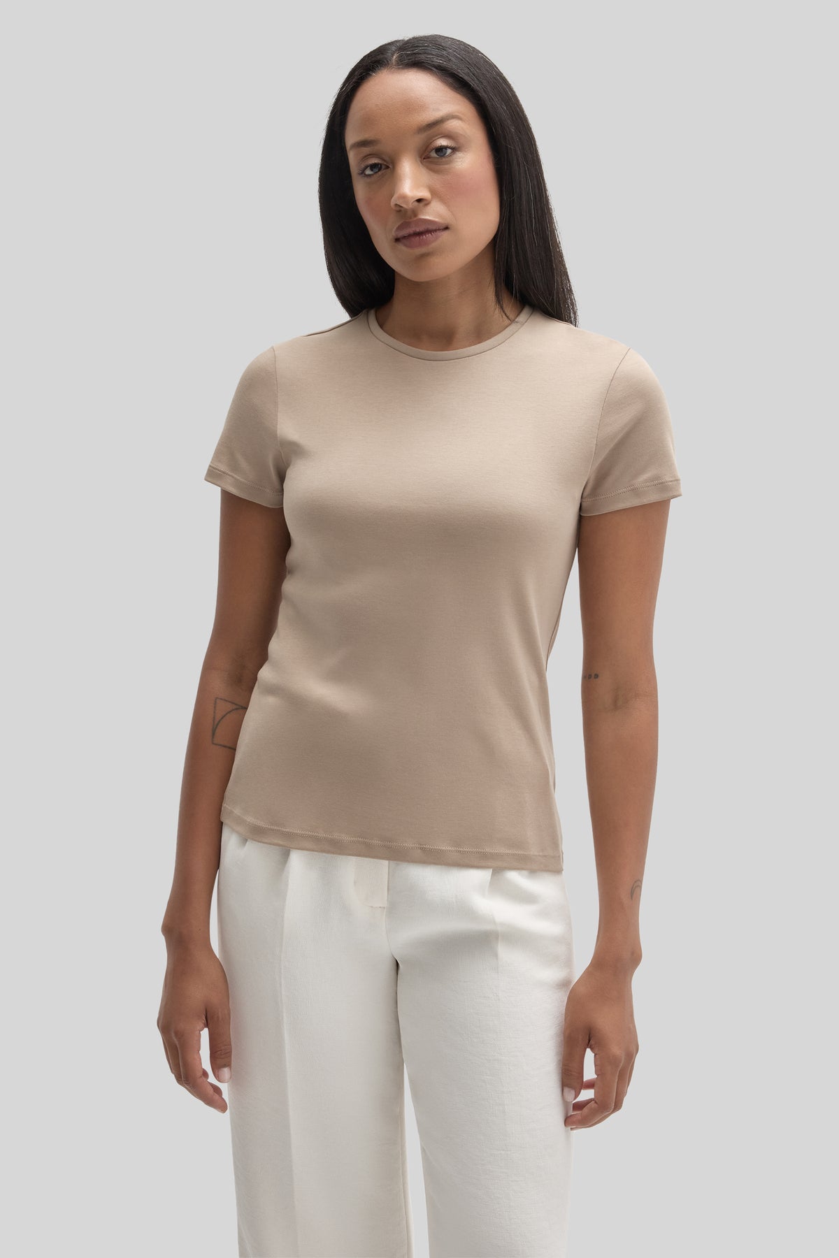 Woman wearing a beige crewneck short-sleeve t-shirt with white pants against a gray background.